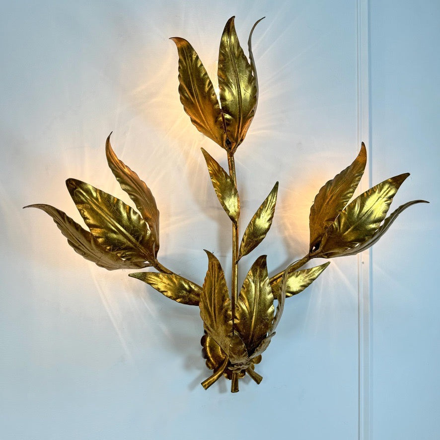 A metallic gold leaf branch wall sconce with multiple leaf-shaped light bulbs, mounted on a light blue wall.
