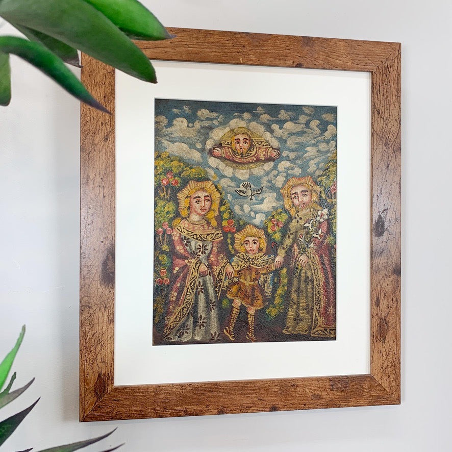 A framed religious cuzco painting depicting the Holy Family (Mary, Joseph, and the child Jesus) under a cloudy sky, housed in a rustic brown wooden frame with a white mat, hanging on a light gray wall with green leaves in the foreground.