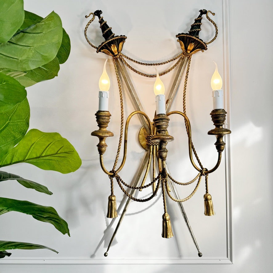 An ornate wall-mounted sconce with three candle-style lights, cross swords, featuring a bronze and gold finish with rope details and tassel accents, shown against a white wall with a green plant leaf on the left.