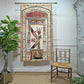 Egyptian-themed wall tapestry applique  hanging on a rod in a styled room, featuring a stylized figure with a feather and hieroglyphic symbols in beige, red, black, and white. A wooden chair and a potted plant are visible in the room.