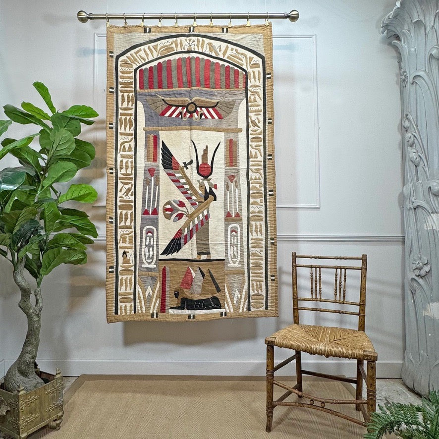 Egyptian-themed wall tapestry applique  hanging on a rod in a styled room, featuring a stylized figure with a feather and hieroglyphic symbols in beige, red, black, and white. A wooden chair and a potted plant are visible in the room.