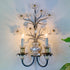 A decorative wall sconce with a dark metal branch structure, crystal flower petals, and two lit candle-style bulbs in clear glass holders, mounted against a white wall.