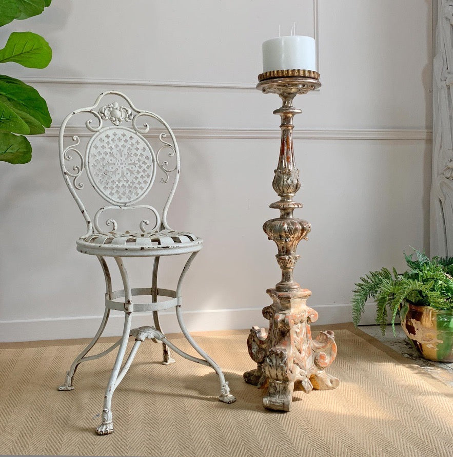 An antique Arras white metal garden chair sits beside a tall, ornate antique  wooden candlestick holder with a white pillar candle, set on a woven beige rug against a white paneled wall.