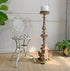 An antique Arras white metal garden chair sits beside a tall, ornate antique  wooden candlestick holder with a white pillar candle, set on a woven beige rug against a white paneled wall.
