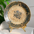 An antique ceramic Safavid plate with a blue floral pattern and crackle glaze, displayed on a gold-colored stand on a white surface with a green plant in the background.