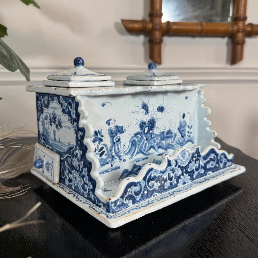 A decorative antique delft blue and white porcelain ink well with two small lidded boxes on top, shown on a dark tabletop in an indoor setting.
