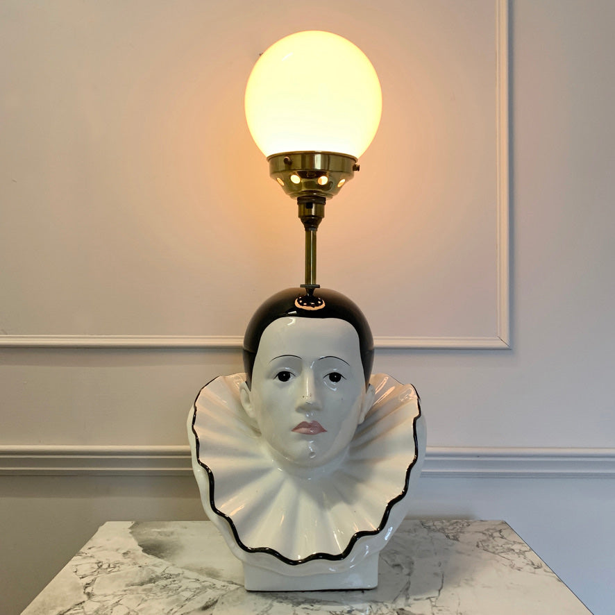 A decorative table lamp shaped like a white porcelain pierrot clown bust with a black ruffled collar, a black hairpiece, and a brass-toned stem supporting a glowing white globe shade, set on a marble surface against a light paneled wall.
