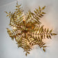 A decorative wall sconce designed to look like metallic gold fern fronds, mounted on a plain white wall with warm light glowing from within.