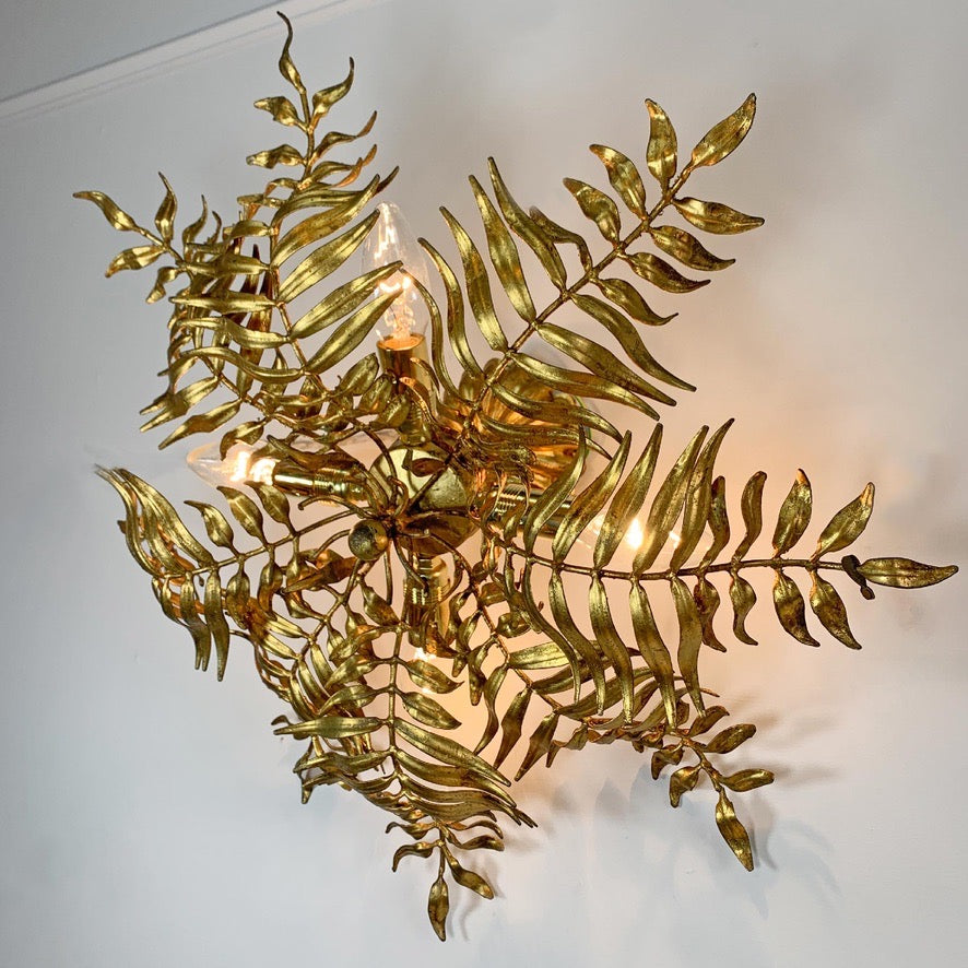 A decorative wall sconce designed to look like metallic gold fern fronds, mounted on a plain white wall with warm light glowing from within.