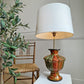 A vintage Kashmiri table lamp with a large white drum shade and a glossy, multicolored ceramic base featuring red, gold, and green tones, styled next to a wooden chair and a potted olive tree.