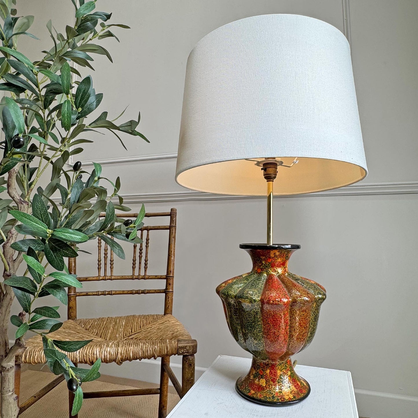 A vintage Kashmiri table lamp with a large white drum shade and a glossy, multicolored ceramic base featuring red, gold, and green tones, styled next to a wooden chair and a potted olive tree.