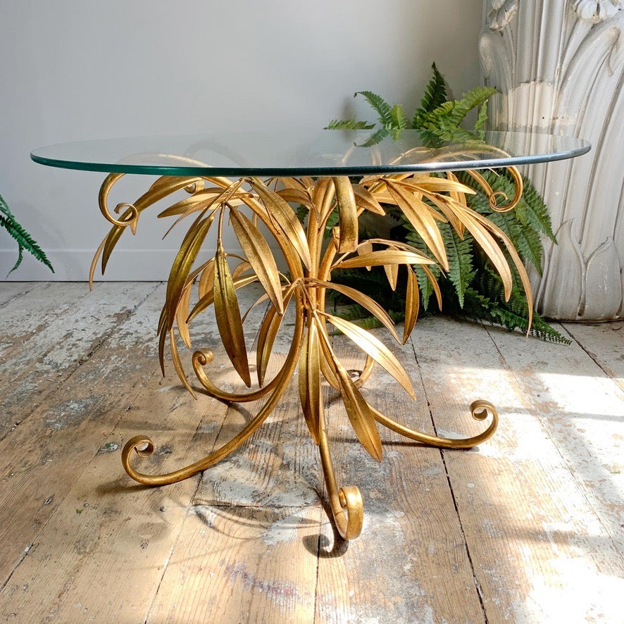 A mid-century modern console table with a gold-painted metal palm-leaf base and a clear glass oval tabletop, set on a rustic wooden floor.