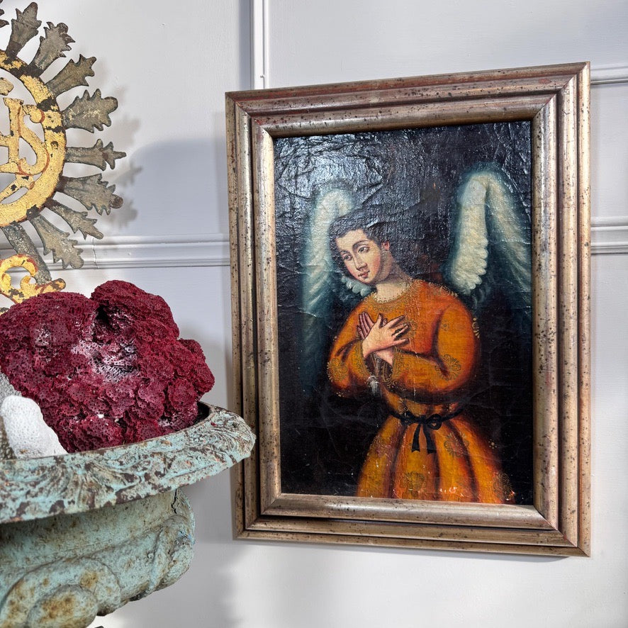A framed religious Cuzco oil painting of an angel with white wings and a brown robe, displayed on a white wall next to a decorative metal sunburst and a stone planter with red flowers.