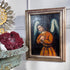 A framed religious Cuzco oil painting of an angel with white wings and a brown robe, displayed on a white wall next to a decorative metal sunburst and a stone planter with red flowers.