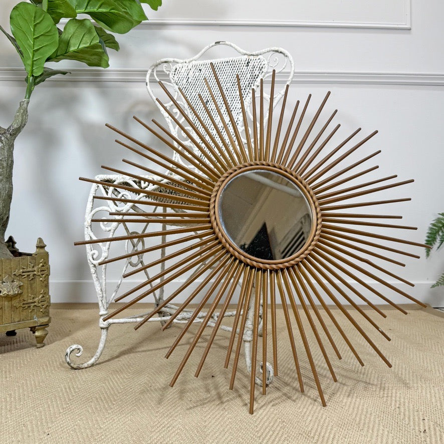 A round Chaty Vallauris sunburst mirror with a natural wood frame and radiating metal rays, displayed in a styled interior setting next to a white wicker chair and a potted plant.
