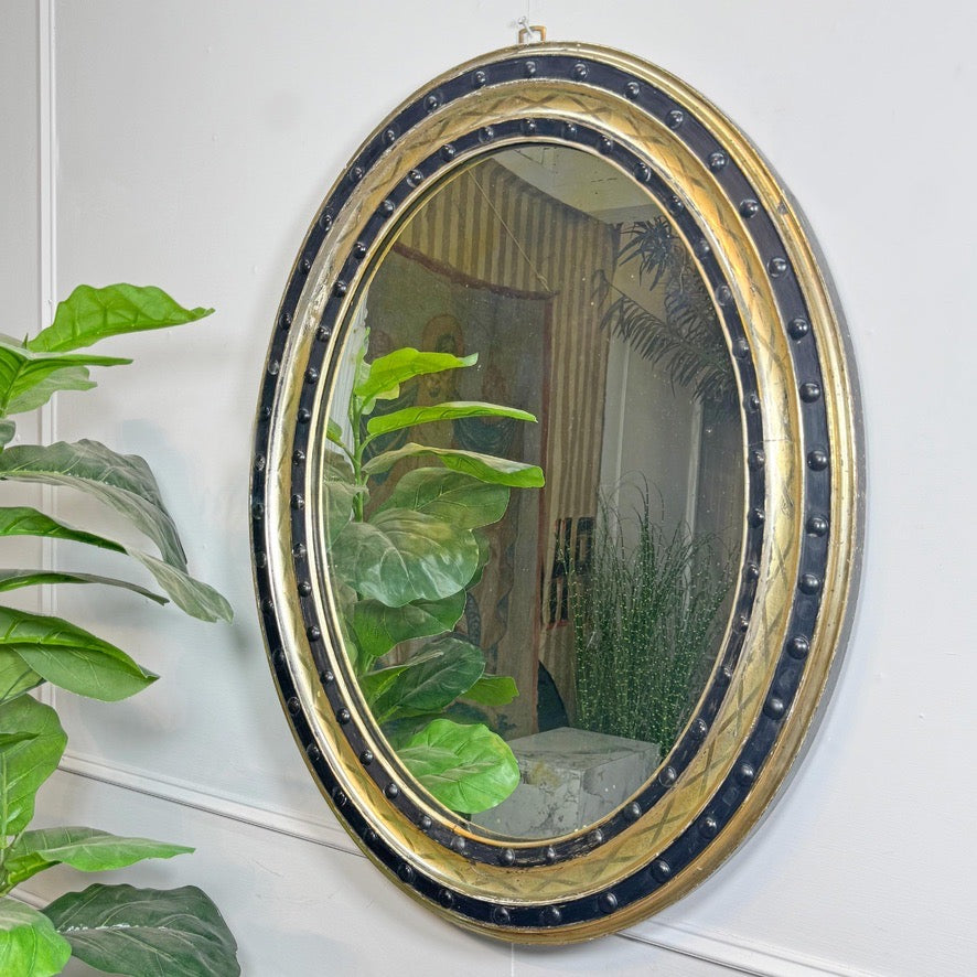 An Irish oval vintage-style wall mirror with an ornate gold frame and a black beaded border, mounted on a white wall with green houseplants visible in the foreground and reflected in the glass.