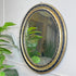 An Irish oval vintage-style wall mirror with an ornate gold frame and a black beaded border, mounted on a white wall with green houseplants visible in the foreground and reflected in the glass.