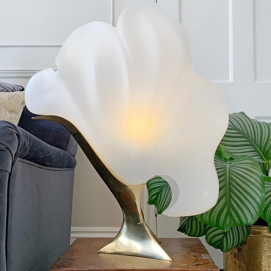 A sculptural table lamp with a large, wavy white frosted shade mounted on a polished gold-toned metal base, styled on a wooden table next to a gray sofa and a green plant.