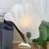 A sculptural table lamp with a large, wavy white frosted shade mounted on a polished gold-toned metal base, styled on a wooden table next to a gray sofa and a green plant.