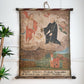 An antique 17th century religious wall scroll painting hanging on a white wall, depicting saints, angels, and a small village scene, with German text at the bottom.