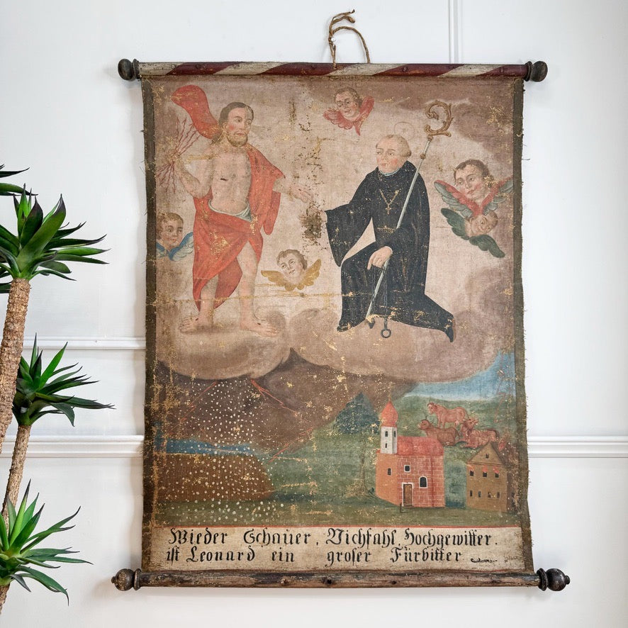 An antique 17th century religious wall scroll painting hanging on a white wall, depicting saints, angels, and a small village scene, with German text at the bottom.