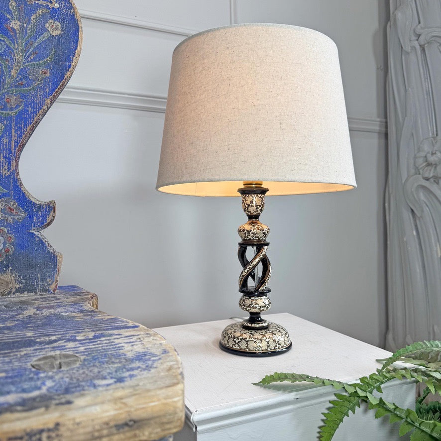 A Kashmiri table lamp with a large beige fabric drum shade and an ornate dark metal base featuring gold and silver mosaic detailing, styled on a white side table next to a blue distressed wooden chair.