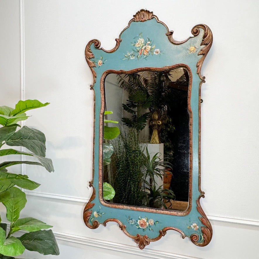 An ornate vintage wall mirror with a distressed turquoise blue frame and carved brown wood details, decorated with delicate floral hand-painted motifs. The mirror is mounted on a white paneled wall next to a green potted plant.