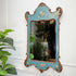 An ornate vintage wall mirror with a distressed turquoise blue frame and carved brown wood details, decorated with delicate floral hand-painted motifs. The mirror is mounted on a white paneled wall next to a green potted plant.