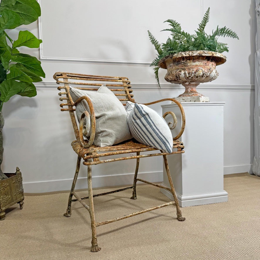 A distressed metal and wicker armchair with two throw pillows, styled in a room setting next to a white pedestal with a large antique cast iron urn planter.