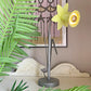 A vintage Bliss table lamp with a gray metal base and neck, featuring a flower-shaped shade that opens into two yellow petals, lit from within. The lamp is styled on a tabletop with green palm leaves in the foreground and a framed art print on a beige wall behind it.