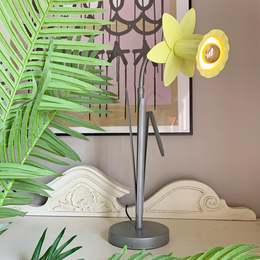 A vintage Bliss table lamp with a gray metal base and neck, featuring a flower-shaped shade that opens into two yellow petals, lit from within. The lamp is styled on a tabletop with green palm leaves in the foreground and a framed art print on a beige wall behind it.
