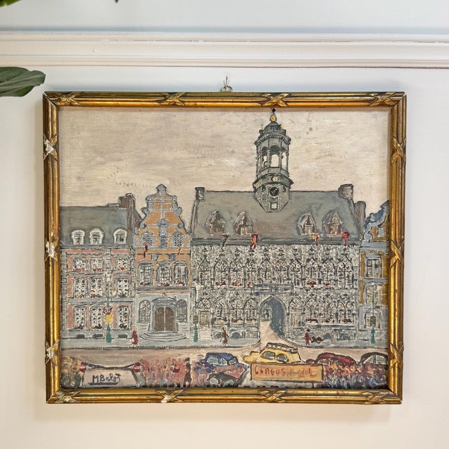 An antique oil painting of a European town square with historic buildings, a clock tower, and street traffic, housed in a distressed gold frame and hanging on a white wall.