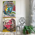 Two colorful abstract portrait paintings leaning against a white paneled wall, with a silver metal chair and a potted fern on a light wood floor.