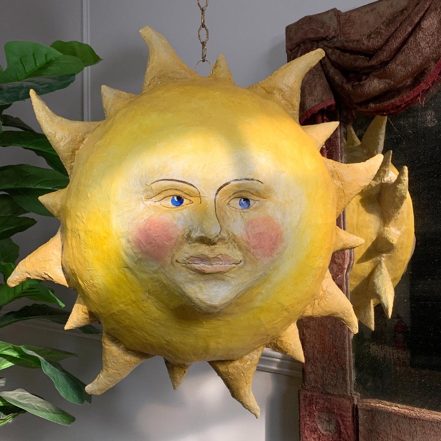 A whimsical hand-painted paper mache yellow sun wall hanging with a smiling face, blue eyes, and pink cheeks, shown hanging in an indoor setting next to a plant and a mirror.
