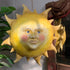 A whimsical hand-painted paper mache yellow sun wall hanging with a smiling face, blue eyes, and pink cheeks, shown hanging in an indoor setting next to a plant and a mirror.