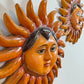A close-up of a handcrafted Sergio Bustamante sun wall hanging with a human face and radiating sun rays, shown against a plain light background. A second similar sun decoration is partially visible in the upper right.