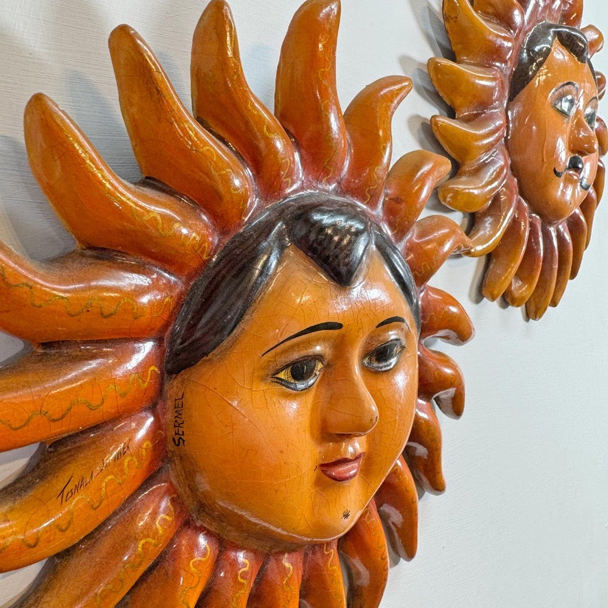 A close-up of a handcrafted Sergio Bustamante sun wall hanging with a human face and radiating sun rays, shown against a plain light background. A second similar sun decoration is partially visible in the upper right.
