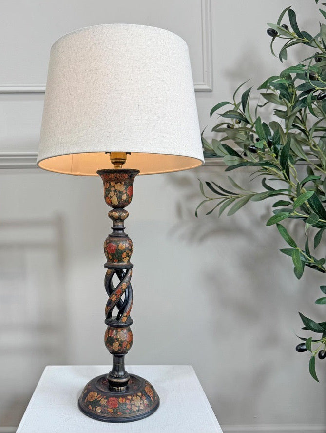 A decorative table lamp with a large off-white fabric drum shade and a dark carved wooden base featuring hand-painted floral details, set on a white pedestal against a light gray paneled wall with a wooden chair and green plant visible.