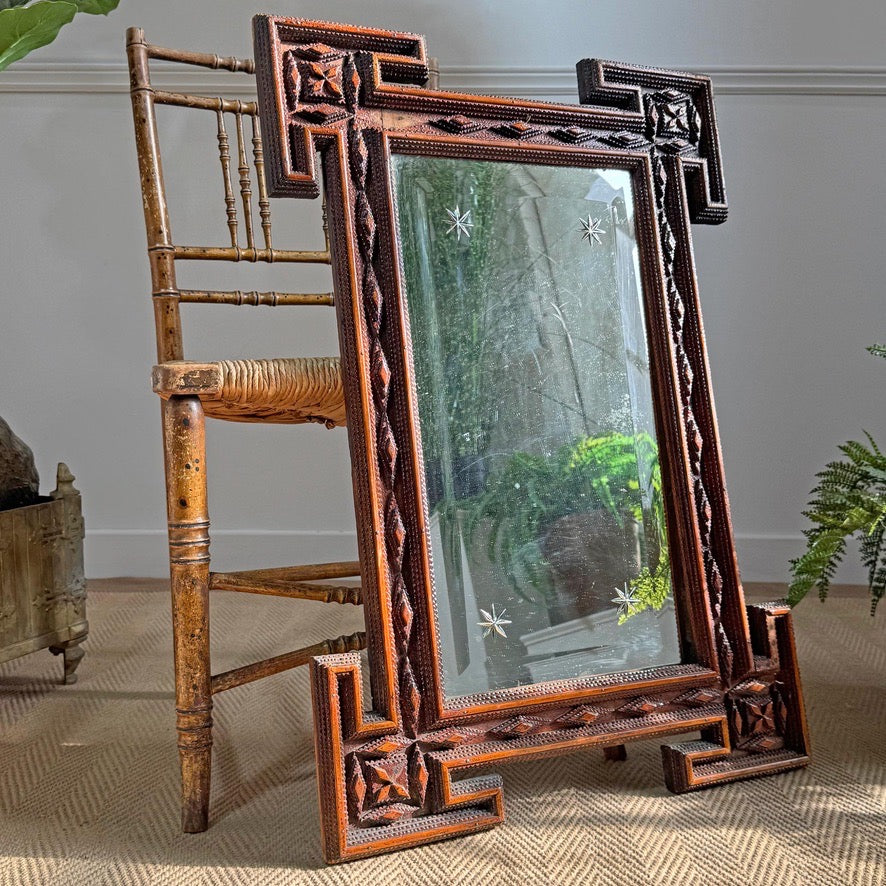 An antique carved Tramp art ornate rectangular floor mirror with a dark carved wooden frame, leaning against a wooden chair on a textured beige rug in an indoor room setting.