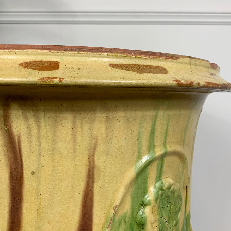 Close-up of a vintage ceramic anduze planter pot with a cream-colored glaze, featuring brown drips and green vertical streaks, with embossed floral details near the base.