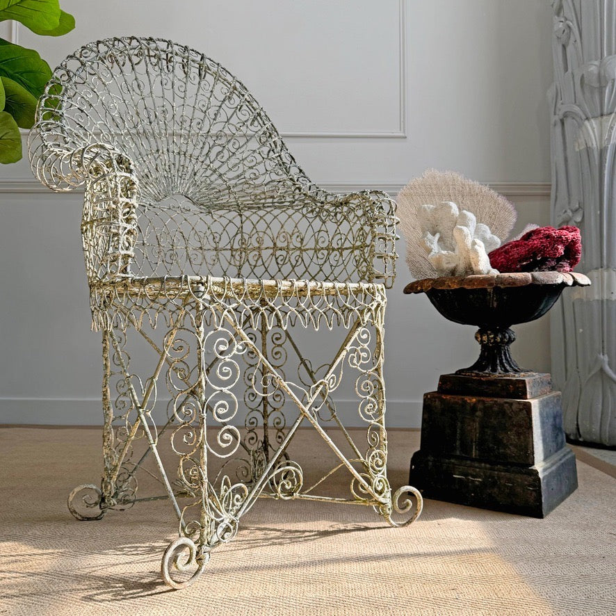 An ornate white antique wirework  metal garden chair with a high back and scrollwork details, shown in a styled interior setting next to a decorative pedestal bowl.