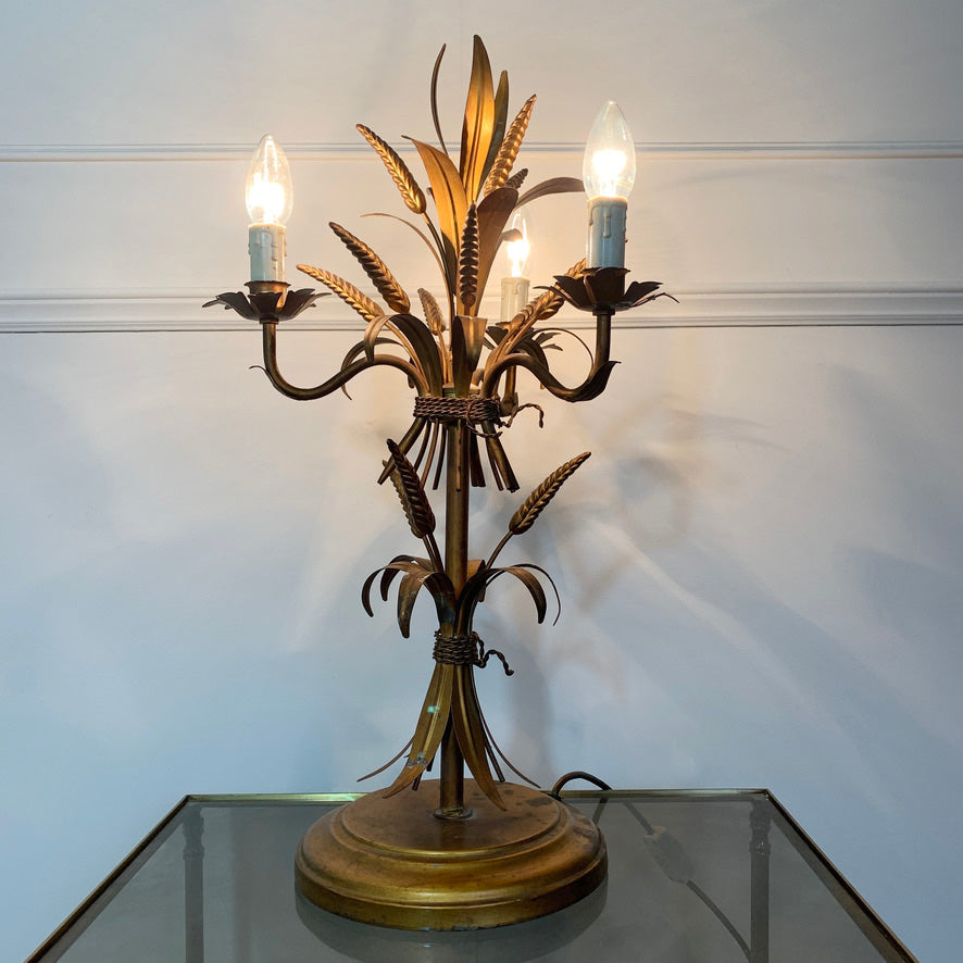 A decorative three-light table lamp with a bronze-finished metal frame shaped like wheat stalks and leaves, shown lit on a glass tabletop against a white paneled wall.