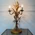 A decorative three-light table lamp with a bronze-finished metal frame shaped like wheat stalks and leaves, shown lit on a glass tabletop against a white paneled wall.