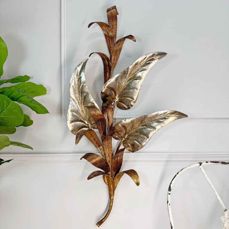 A decorative metal wall hanging shaped like a leafy plant stem, with metallic gold and bronze-toned leaves mounted on a white paneled wall. A green plant leaf appears at the left edge, and part of a white chair is visible at the lower right.