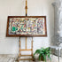 A framed abstract painting with stylized cat figures, displayed on a wooden easel in a bright room with a white wall, gray chair, potted fern, and decorative column.