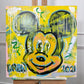 A square canvas painting on an easel featuring a stylized Mickey Mouse face in yellow, black, green, and blue with dripped paint effects and the text 'BREW 2021' on a light gray wall background.