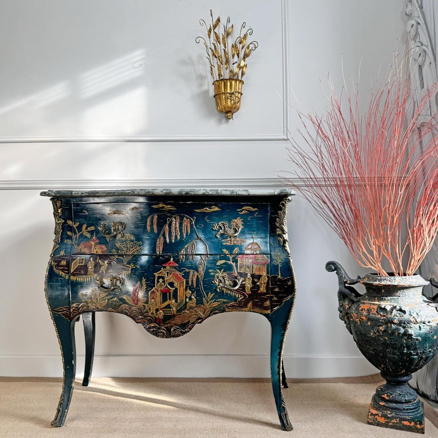 A decorative antique blue commode set of drawers with an ornate multicolor chinoiserie landscape design, set against a white panelled wall next to a large decorative urn with pink branches.