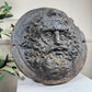 A round, dark gray cast-iron relief wall plaque featuring a carved face of Zeus  with long curly hair and a beard, displayed on a white surface with a light-colored paneled wall and green leaves in the background.