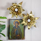 Two gold-toned maison jansen metal flower-shaped wall sconces with white globe bulbs, displayed above a small religious icon on a white wall, with green leaves entering from the left.