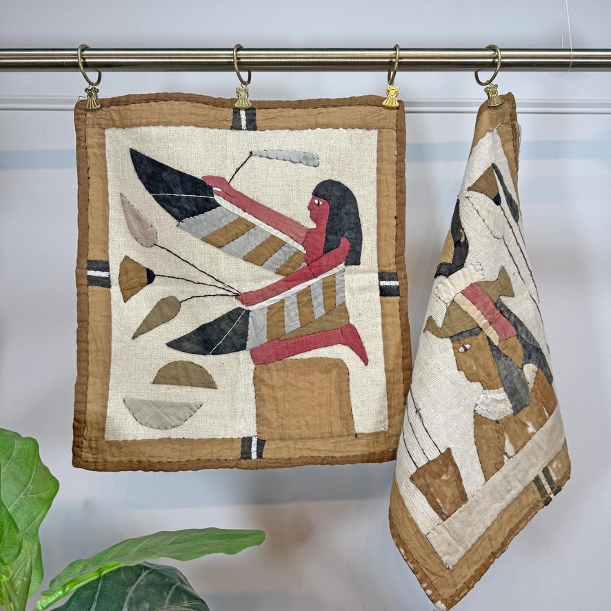 Two vintage Egyptian-style quilted cloth hangings from gold-colored hooks on a metal rod. The napkin on the left is fully displayed, showing a figure with a feathered headdress in beige, brown, black, red, and white.